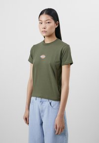 Olive green short-sleeve t-shirt with a round neckline, featuring a circular logo on the chest. Paired with light blue relaxed-fit jeans.