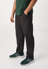 Pantalons cargo noirs à coupe décontractée, avec des poches latérales et une texture de tissu lisse. Assortis à des baskets vertes avec des accents blancs.