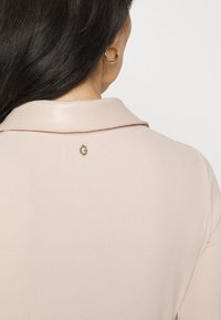 Femme aux longs cheveux foncés portant un t-shirt rose clair côtelé avec un col en cuir et un petit emblème doré en forme de "G" dans le dos.