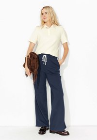 T-shirt polo crème, pants de jogging navy à jambes larges avec cordon blanc, tenant un sac à franges marron, se tenant contre un fond blanc.