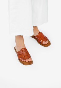 Sandali in pelle marrone con design a strisce intrecciate e suola piatta, abbinati a pantaloni bianchi a gamba larga, con le dita dei piedi che spuntano.