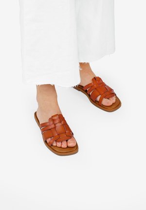 Sandali in pelle marrone con design a strisce intrecciate e suola piatta, abbinati a pantaloni bianchi a gamba larga, con le dita dei piedi che spuntano.
