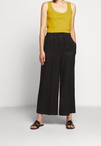 Mustat löysät housut, joissa on taskut ja kiristysnauha vyötäröllä, yhdistettynä sinapinkeltaiseen hihattomaan paitaan. Kangas näyttää pehmeältä ja siinä on hieman kiiltoa.