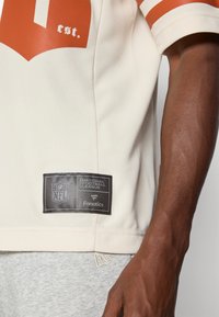 NFL dres s krémovou tkaninou, oranžovými akcenty a síťovanou texturou. Obsahuje černou nášivku loga NFL a branding "Fanatics" na lemu.