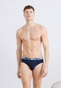 Άνδρας χωρίς πουκάμισο, που φοράει σκούρα μπλε εσώρουχα με επώνυμη μέση, στέκεται μπροστά σε απλό ανοιχτό φόντο.