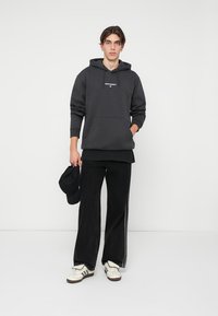 Sudadera gris con capucha y bolsillo frontal, acompañada de jeans negros de pierna ancha y zapatillas blancas con detalles en negro.