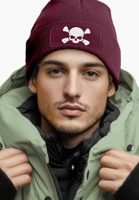 Maroon beanie with skull and crossbones patch, ribbed texture. Green parka with black lining, fur-collared, tightly fitted around the face.