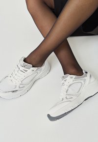 Bílé sportovní tenisky s prodyšnými a syntetickými překryvy, texturovanou podrážkou a šedými akcenty, v kombinaci s průhlednými černými punčochami.