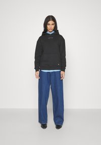 Kvinne iført en svart Tommy Jeans hettegenser over en blå skjorte, brede blå jeans og svarte spissede sko, som står mot en enkel hvit bakgrunn.