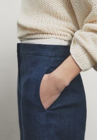 Persoon met hand in de zak, gekleed in marineblauwe broek en een crèmekleurige gebreide trui met opgerolde mouwen, weergegeven vanaf de taille naar beneden.