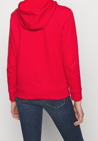 Červená mikina s kapucí na stažení, hladká textura, žebrované manžety a lem, v kombinaci s tmavě modrými džínami. Pohled zezadu neukazuje žádné viditelné vzory.