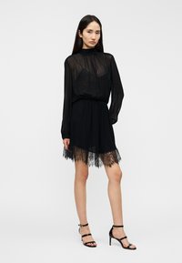 Robe noire avec manches longues transparentes, taille froncée et bordure en dentelle à l'ourlet. Texture lisse et tissu léger pour un aspect fluide.