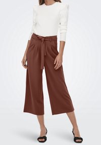 Pantalon large marron à taille ceinturée, associé à un haut blanc côtelé avec des manches bouffantes. Le mannequin porte des sandales à talons noires.