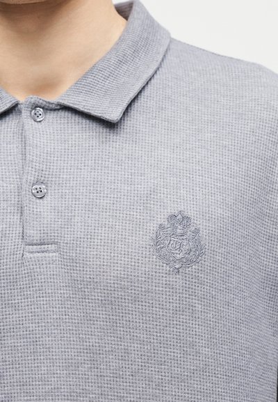 Κοντινή λήψη ανοιχτόγκρι πλεκτού πόλο πουκάμισου με δύο κουμπιά και κεντημένο έμβλημα με το οικόσημο στο αριστερό μέρος του στήθους.