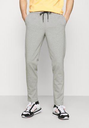 Mann trägt hellgraue Jogginghose mit schwarzem Kordelzug und weiße, schwarze und rote Turnschuhe, steht mit den Händen in den Taschen.