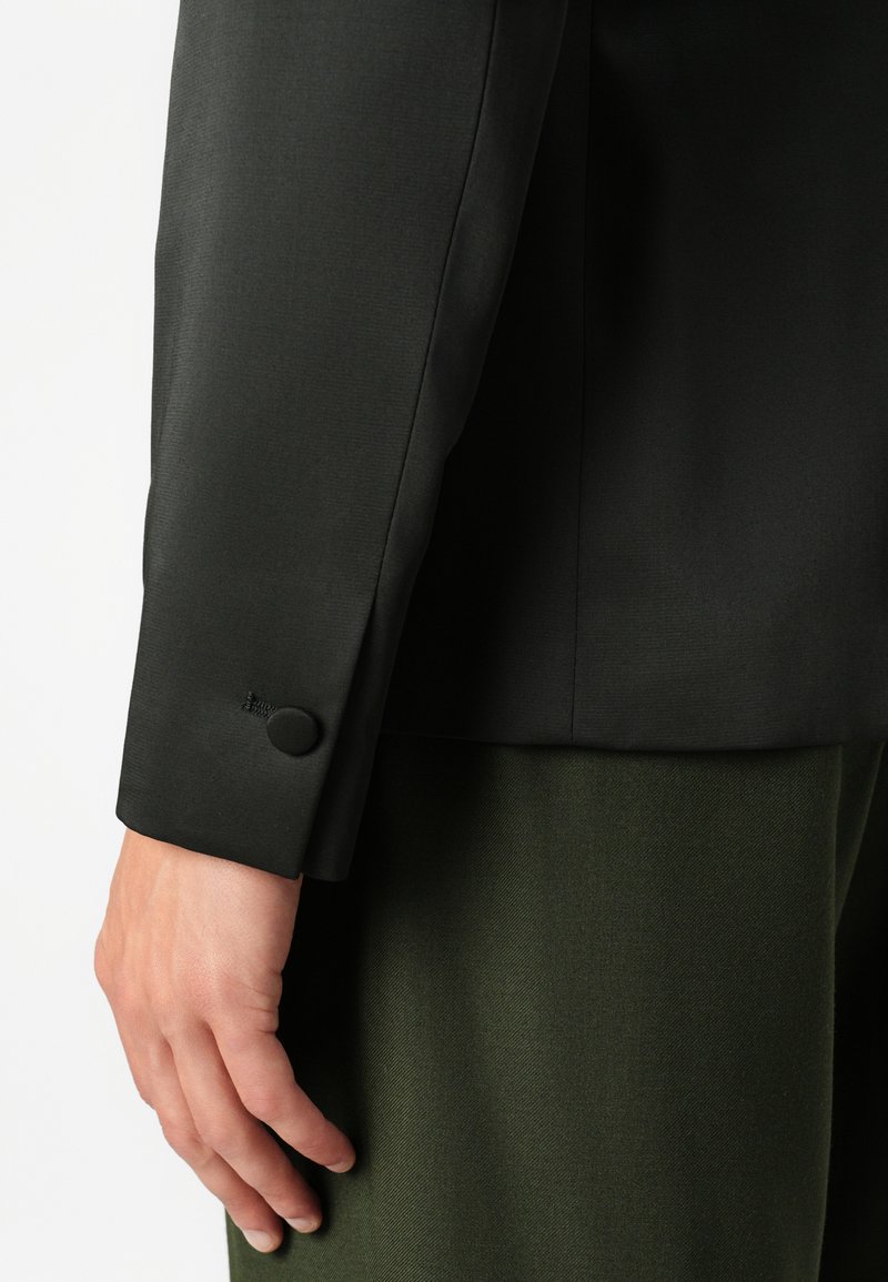 Manche de blazer noir ajusté avec un détail de bouton, complétée par un pantalon texturé vert foncé. Finition en tissu lisse sur la veste.