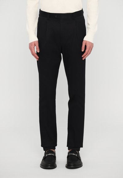 Pantalons noirs ajustés avec un devant plat, texture minimale et une ceinture. Associés à des chaussures noires avec une chaîne argentée décorative.
