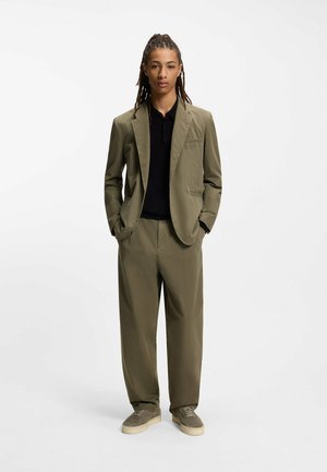 Olive green suit with a tailored blazer and loose-fit trousers. Black polo shirt underneath. Gray sneakers complement the outfit.
