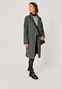 Manteau à motifs gris avec un col, des manches longues et une fermeture à boutons à l'avant. Porté sur un haut gris avec une jupe noire. Bottines noires à la cheville et sac à bandoulière.