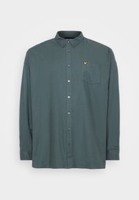 Camisa de botones de manga larga en tejido verde oscuro, con un bolsillo en el pecho y un pequeño logo amarillo en el lado izquierdo. Textura suave.