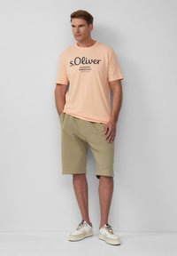 Pfirsichfarbendes T-Shirt mit Rundhalsausschnitt und schwarzem Druck, kombiniert mit hellgrünen Shorts und weißen Sneakers, die beigefarbene Akzente aufweisen und einen lockeren Schnitt haben.