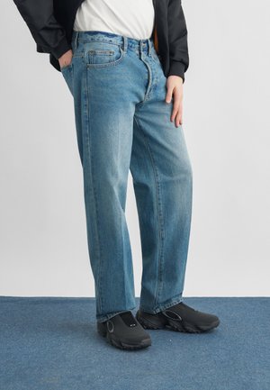 Personne portant un jean bleu ample, des chaussures noires sans lacets, une chemise blanche et une veste noire, debout sur un tapis bleu.