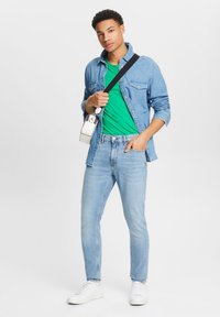 Blugi deschiși, un tricou verde și o cămașă din denim albastru, asortate cu adidași albi. Poartă o geantă mică albă.