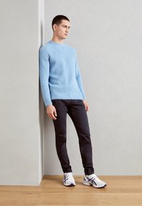 Pullover lavorato a maglia azzurro chiaro, con polsini e orlo a coste, abbinato a pantaloni scuri e sneakers bianche con accenti viola. In piedi contro un muro.