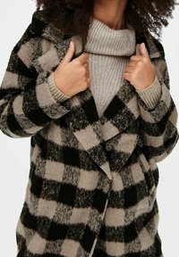 Manteau à carreaux noir et beige, fabriqué en mat ériau texturé, présentant une coupe croisée et un grand col à rabat.