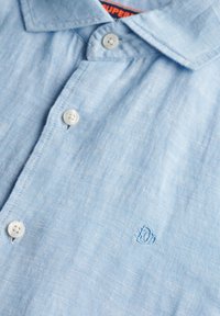 Lichtblauw linnen shirt met een button-down kraag, voorzien van twee witte knopen en een subtiel geborduurd logo op de borst.