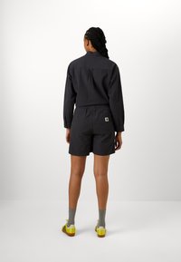 Zwart geruit overhemd van lichtgewicht stof, korte broek met elastische tailleband, grijze sokken en schoenen met gele accenten. Achteraanzicht.
