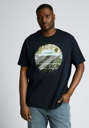 Mann trägt schwarzes T-Shirt mit Vintage-Auto-Grafik und Text, steht mit einer Hand in der Tasche vor einfarbigem Hintergrund.