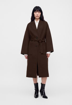 Un mannequin se tient devant un fond uni, portant un manteau marron foncé ceinturé, longueur genou, une chemise blanche et des bottines noires à talons.