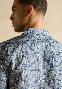 Modrá košeľa s kvetinovým vzorom, ktorá sa vyznačuje podrobným bielym kvetinovým dizajnom, krátkym golierom a bezpečným zapínaním s dvoma viditeľnými gombíkmi na zadnej strane.