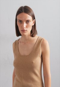 Top sans manches beige en tissu tricoté doux, avec un décolleté arrondi, une texture lisse et des coutures minimales pour un look épuré.
