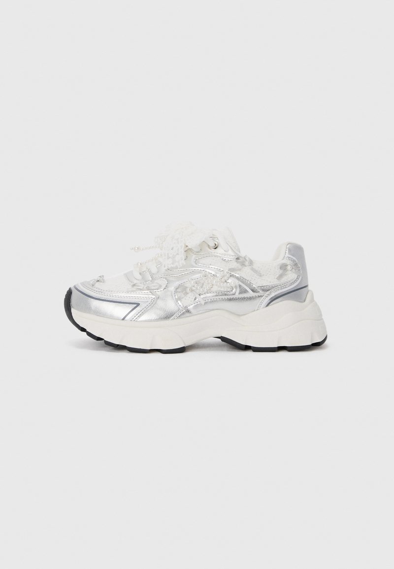 Baskets de sport argentées avec une tige texturée, lacets blancs, semelle noire contrastante et design courbé. Accents métalliques remarquables sur les côtés.