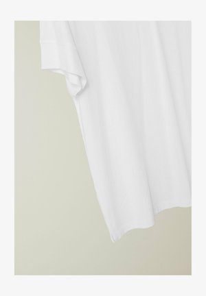 Camiseta blanca de algodón de manga corta colgada contra una pared beige claro, mostrando detalles de la parte superior y la manga.