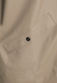 Beiger wasserdichter Stoff mit glatter Textur, ausgestattet mit einer Klapptasche, die mit einem Druckknopf gesichert ist, und kontrastierenden Nähdetails.
