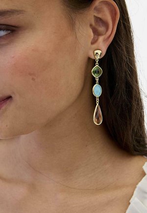 Mujer con un pendiente colgante de oro con tres piedras de colores: cuadrado verde, óvalo azul y formas de lágrima ámbar.