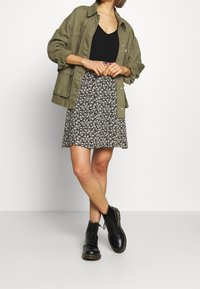 Khaki oversized jasje over een zwarte getailleerde top, gecombineerd met een zwarte rok met bloemenprint en zwarte veterschoenen, op een neutrale achtergrond.