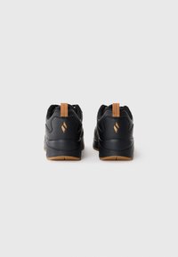 Black athletic shoes with leather and mesh materials, prominent rubber soles, and tan accents at the heel. Minimalist design, rear view.