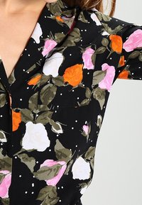 Blouse noire avec un motif floral présentant des roses en rose, blanc et orange. Tissu lisse avec un col et des détails de boutons.