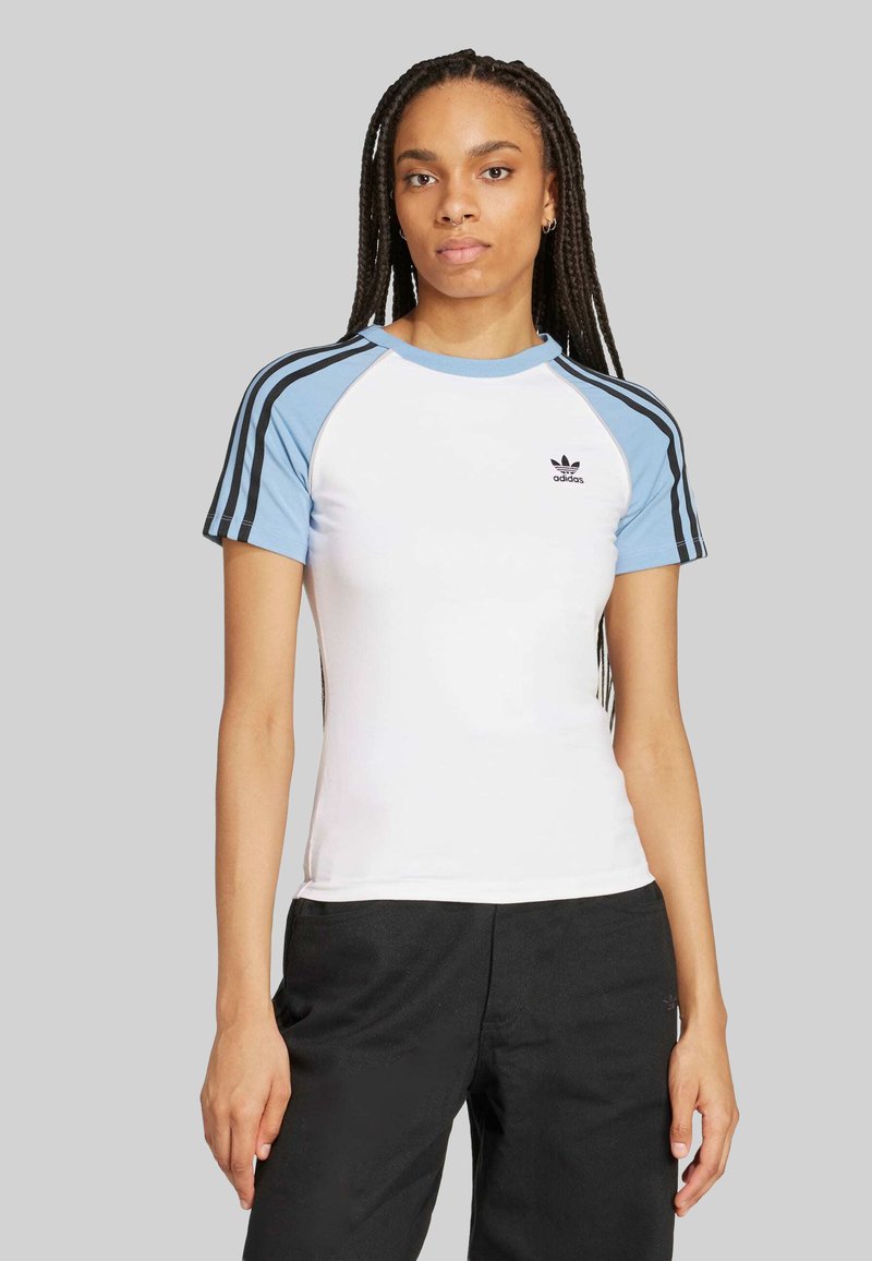 Bijela majica s kratkim rukavima koja dobro pristaje, s svijetloplavim raglan rukavima, crnim trostrukim detaljima i malim crnim logom na prsima. Glatka tekstura tkanine.