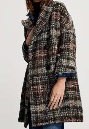 Femme portant un manteau épais en laine à carreaux dans des tons noir, gris, blanc et rouge avec de gros boutons noirs et un revers en jean bleu en dessous.