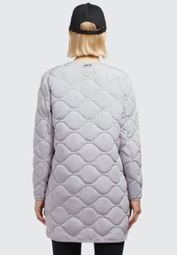 Manteau long matelassé gris clair avec un motif ondulé et un col rond, arborant une casquette noire portée à l'envers. Accents noirs sur les manches.