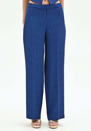 Pantalon large bleu à fines rayures verticales portées avec des sandales à plateforme ouvertes dorées sur un fond uni.