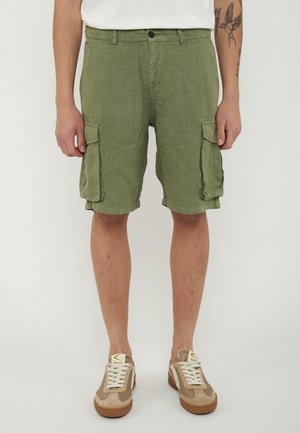 Uomo che indossa pantaloncini cargo verdi con tasche laterali, sneakers beige e bianche, mostrando gambe e braccia tatuate su sfondo bianco.