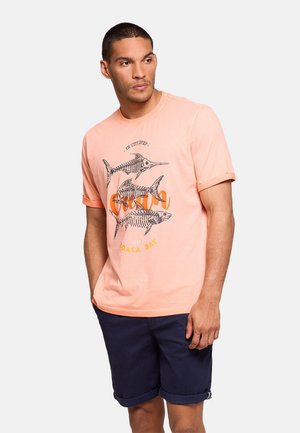 Hombre con camiseta color melocotón con gráfico de esqueleto de pez y pantalones cortos azul marino, de pie con las manos en los bolsillos, mirando hacia un lado sobre un fondo liso.