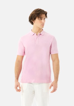 Mand iført en lyserød kortærmet polo og hvide bukser, kigger mod højre mod en ensartet lys baggrund.