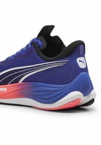 Zapatilla de running morada con parte superior de malla texturizada, acentos en negro, entresuela en rosa y blanco, y un logo distintivo en el talón.